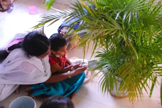 A candid photo taken from above of a woman in a white sari with a long braid sitting on a tiled floor beside a potted palm plant. She leans over a young child in a red top and blue skirt, and both hold a small object between their hands. Another person in blue sits partly visible at the top, and a child's head is visible at the bottom edge near a plastic container. The scene appears to be an indoor gathering with the palm fronds prominent in the foreground.