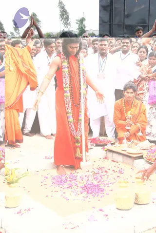 A photograph of an outdoor ritual on a sandy ground, with a long white pole or post set upright in the centre. A barefoot man in a red sleeveless top and red-orange dhoti stands beside it, wearing a long garland of white, purple and yellow flowers and a rudraksha mala, with a gold bangle on one wrist. Scattered pink and purple flower petals cover the sand. Behind him a crowd of men in white shirts and dhotis, some with lanyards, and women in patterned saris stand with palms pressed together in namaskar. On the right, a young man in orange robes with a flower garland kneels beside a tray of petals and a small oil lamp, and a brass kalasha-style vessel sits nearby. Two woven baskets hold yellow flowers in the foreground. A figure in orange at the left raises one arm.