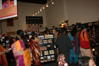 A busy indoor bookshop-style gallery with white walls and track lighting. A woman in the foreground wears a yellow and magenta sari with her long dark hair in a braid. Beside her, a woman in a red and gold sari stands in the crowd. At right, a woman in an orange and red top and a man in a dark jacket browse shelves. A wall poster reads Life Bliss Meditation and shows people seated in meditation poses. A flat-screen displays a Living Enlightenment advertisement. Shelves hold books and CDs displayed on racks, framed photographs line the back wall, and a small brass figure with a red and white flower garland sits in the lower right corner.
