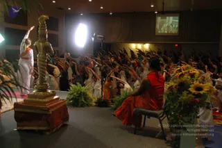 A yoga session in a large hall with a crowd of participants in white and light clothing, arms raised overhead in a stretch. In the foreground, a seated figure in a red-orange robe sits on a folding chair facing the group, with a red-orange wrap and long dark hair in a braid. A tall golden lamp stand with a spiral shaft stands on a red-draped pedestal on the left, and a standing woman in white with an orange sash raises one arm near it. Bouquets of sunflowers and red flowers and green ferns decorate the stage. A projection screen and stage lights are visible at the back.