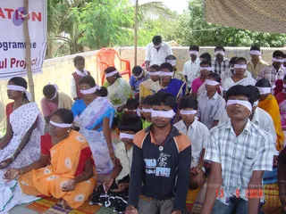 A photograph of an outdoor meditation session with many seated participants, all wearing white cloth blindfolds over their eyes. They sit on woven mats under a shaded canopy beside a low wall and green trees. In the foreground a young man in a grey-and-navy sweatshirt and a man in a checked short-sleeved shirt sit cross-legged with hands resting on their knees. Women in saris, one in orange with a red blouse and another in blue, sit among them, and a man in white stands at the back. A partially visible white banner at left reads 'ation' and 'gramme' and 'ual Project'. A date stamp in the lower right reads 13 9 2009.
