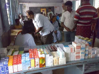 A photograph of a free medical camp held in a classroom with a blue-grey wall and a chalkboard. A man in a white shirt and grey trousers leans over a long blue table stacked with medicine boxes, bottles and tins, while a woman in a white coat with an orange-edged garment sits at the table in the foreground. Several villagers stand and sit along the room, including an older man in white, a woman in a pale blue sari, and a man in a red striped shirt standing at right. A framed picture hangs on the wall above the chalkboard, which bears a handwritten date.