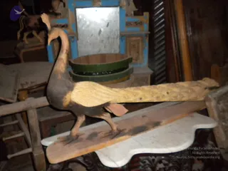 A photograph of a carved wooden peacock sculpture standing on a long wooden plank placed over a white marble slab. The peacock has a pale yellow-tan tail fanned out behind it and a dark brown-grey body and neck, with a raised crest on its head. Behind it sits a wooden tub with green and black painted bands, in front of a blue-painted wooden cabinet with a mirror. The setting looks like a cluttered display room with other wooden objects and furniture.