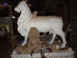 A large white stone sculpture of a seated bull, Nandi, with carved harness, bells, tassels and a decorated saddle cloth marked with a central motif, stands in a dim workshop. Beside it, an unfinished brown stone figure of a multi-armed deity murti, carved with a crown-like headdress and holding a long implement, sits on a white pedestal. Workshop tools, a bucket, a bicycle and stacked materials are visible, along with framed images on the left. No people are visible.