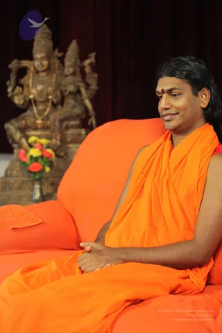 A seated man in an orange saffron wrap sits on an orange-upholstered sofa, hands clasped in his lap, with a red bindi on his forehead and long dark hair swept back. He wears an orange garment over a red top and looks off to the side with a slight smile. Behind him stands a bronze-coloured deity murti of a standing couple with a gold garland, beside a vase of yellow and red flowers against dark red drapery. A small orange cloth rests on the armrest. The man has long swept-back hair, orange saffron clothing and a red bindi, which are the qualifying cues for a probable Swamiji, seated in a ritual-room setting.