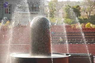 A photograph of a large dark cylindrical lingam, a Shiva linga, standing on a round basin while water sprays and cascades from jets around it. Behind it, tiered red-brick steps hold rows of small potted plants with yellow flowers, and a grey building and trees rise in the background. A few tiny figures, a worker in light clothes near the top of the steps and a person seated lower down in yellow, are visible but are small and partly obscured. The scene is a sunlit outdoor setting with water mist catching the light.