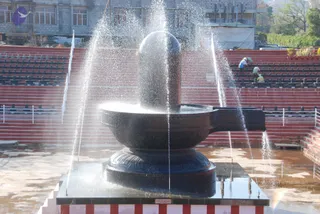 A photograph of a large black stone Shivling standing on a circular yoni pedestal in an open outdoor courtyard, with water jets spraying up around it from the sides, creating a mist. Red-painted tiered seating stands behind it, and a few workers in the background tend to the area beside a multi-storey concrete building. A small plaque sits on the base of the platform. No people are prominently in the foreground, and no one is shown in ritual activity at the Shivling itself.
