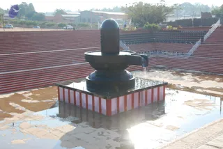 A large black stone Shiva lingam stands on a square red-and-white striped plinth in an open outdoor courtyard, with its yoni-shaped pedestal and spout water channel running out to the side. Water flows from the spout and the stone surrounding area is wet with reflections of the sky. Behind it, tiered red-brick steps rise in a semicircular amphitheatre with white metal railings and stairs, and a few small vehicles and low buildings with trees sit in the background under a hazy morning sky. No people are visible in the scene.
