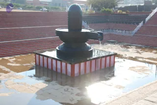 A large dark stone Shiva lingam with a rounded top and a spout-like yoni pedestal stands on a square base striped red and white, set in a shallow pool of water on a wet paved court. Red-brown tiered stone steps with white railings rise behind it, and a few parked vehicles and low buildings appear in the distance under bright sunlight.
