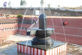 A large black stone Shivling stands on a square platform with red and white striped sides, with water jets spraying up around it from the base. Behind it, a red-brick tiered amphitheatre with stone steps and rows of potted plants rises toward a fenced hillside. Two or three people in red and pink saris stand on the steps at the right, and a few others are near the left edge. The ground is wet from the water.