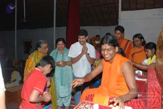 A photograph of an indoor hall with a thatched roof and white walls. In the foreground, a smiling man with long dark hair wears an orange sleeveless top and orange shawl, seated on a red patterned mat and reaching toward a red tray with small items. Facing him, a young boy in a red shirt bends forward with hands together. Behind them, a woman in a yellow sari and glasses stands beside a woman in a teal salwar kameez with a teal shawl holding a small item, and a man in a white shirt presses his palms together. Further back, people in orange clothing and a girl in yellow stand near a red curtain, with a white-clad figure seated at the left edge.