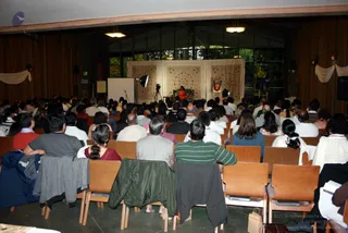Photograph from the back of a wood-panelled hall where a large audience sits on wooden chairs facing a stage. On the stage, a figure in orange clothing sits on a seat in front of a draped cream and patterned backdrop, with a framed picture beside it. A video camera on a tripod and a studio light are set up near the stage, and a white presentation board stands to the left. Many attendees are seen from behind, some in jackets, sweaters and kurtas, and jackets are draped over several chairs in the foreground. Decorative white swags with small floral accents hang from the side walls under tall windows showing trees outside.
