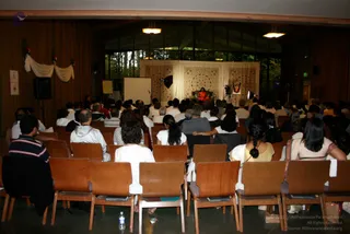 Photograph of a seated audience in a wood-panelled hall facing a stage at the far end. A figure in orange robes sits on a chair in front of a draped backdrop of white and cream embroidered cloth, with a framed picture beside it. Rows of wooden chairs hold many attendees, mostly seen from behind, wearing white, yellow and striped clothing. Large windows show trees outside, and a white projection screen and studio lights stand near the stage. A water bottle lies on the floor in the foreground.