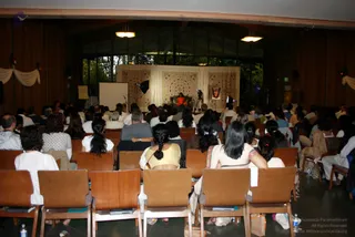 A photograph taken from the back of a wood-panelled hall filled with rows of wooden chairs and many seated attendees, mostly seen from behind. At the far end, a figure in orange clothing sits on a raised chair before a backdrop of cream draped fabric with embroidered patterns. A projection screen stands to the left, and a framed portrait hangs beside the stage on the right. Several attendees wear white and pale outfits, and one woman in the foreground has a long braid. Hanging cream drapes decorate the upper corners of the room.