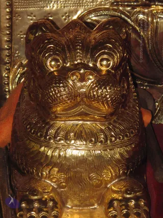Close-up photograph of a gilded lion-shaped sculpture. Behind it are embossed gold panels with dotted borders and floral motifs, and a sliver of orange cloth appears at the left edge. No people are visible; the image shows only the ornamental gold metalwork.
