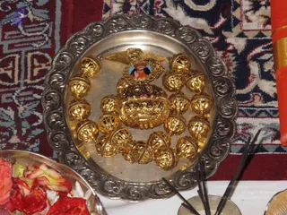 Overhead photo of a silver tray with a repoussé scalloped rim holding a set of ornate gold objects arranged in a circle: a gold lidded bowl-like form with ruby-coloured stones and a gold triangular finial, surrounded by large openwork gold beads. A small framed printed photo of a person in orange clothing with a red head covering is set into the gold piece. Rose petals sit in a silver bowl at the lower left, incense sticks lie across the lower right, and the tray rests on a red and blue patterned rug with an orange-and-gold cloth at the upper right. No people are visible.