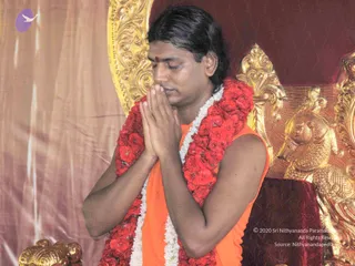 A man with long dark hair sits with eyes closed and palms pressed together in namaskar before his chest. He wears an orange sleeveless garment and a red and white flower garland around his neck, with a red tilak mark on his forehead. Behind him is an ornate golden throne with carved floral and scrollwork designs against deep red fabric, and a draped pinkish-brown curtain stands at left. The scene appears to be a ritual setting with a seated figure in devotion.