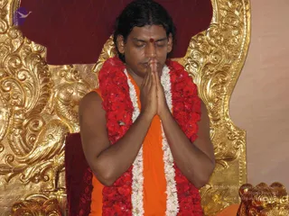 A man seated on an ornate gold throne with carved floral and bird motifs and a deep red cushioned backrest, palms pressed together in anjali mudra with eyes closed. He wears an orange saffron garment, a red flower garland and a white flower garland draped over his shoulders, and a red bindi on his forehead. His hair is long and swept back. The setting suggests a seated honouring or pada puja occasion, though the page gives no visible confirmation of the event itself.