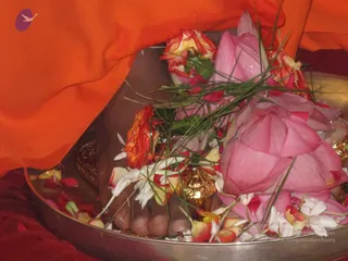 Close-up photo of a pair of bare feet resting in a shallow round metal tray during a pada puja. The feet are surrounded by pink lotus blossoms, red and orange roses, white jasmine-like flowers, scattered petals, and long green grass blades. A small ornate brass vessel stands near the feet, and an orange cloth drapes over the upper edge of the frame, with a red cloth below. No other people are visible.