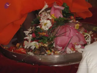 Close photo of a large shallow metal tray filled with flower offerings: a pink lotus bloom, white chrysanthemum-like flowers, white jasmine-type blossoms, yellow petals, red petals and green leaves with grass stems. Orange fabric draped over the tray at top and a white cloth at right edge. Pale rice-like grains are scattered on the tray and a small brass-coloured item is partly visible among the flowers.