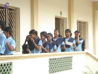 A photograph of a group of schoolchildren in light blue uniform shirts and navy blue pinafore-style vests standing in a line along a covered veranda of a cream-coloured building. Several girls with braided hair, some wearing spectacles, hold small white slips of paper, and a boy in a blue shirt stands at the left edge. Behind them are barred windows and a door marked with the number 3, and a patterned green concrete screen wall runs along the bottom of the frame.