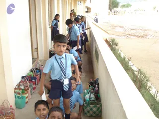 A photograph of a long covered school corridor with students in light blue shirts and navy shorts or skirts, many wearing striped ties and white ID badges on lanyards. A boy in the foreground stands closest to the camera, while others sit and line up along the walkway. Woven baskets, bags and water bottles are placed along the wall. No a person candidate is visible in the scene.