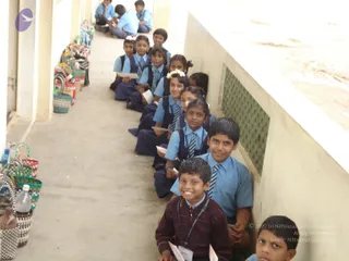 A photograph of schoolchildren in blue shirts, navy trousers and skirts, and striped ties sitting in a row along a concrete covered walkway. Several children hold small white papers and wear lanyards with name badges. A girl in front wears a white flower garland in her hair. In the foreground, two boys smile at the camera, one in a maroon sweater with a lanyard. To the left, many woven baskets and bags with colourful handles are lined up against the wall. Plastic water bottles sit among the baskets. A green lattice panel is visible on the right wall. No adult or ritual figure is visible.