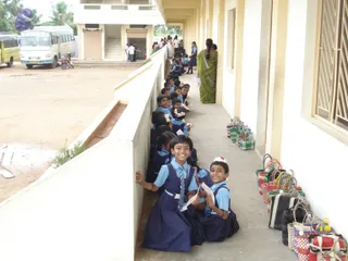 A photograph of schoolchildren seated in a long line along a covered concrete corridor beside a whitewashed wall. Two girls in the foreground wear blue short-sleeved shirts, navy pinafore-style dresses and striped ties; one smiles at the camera and holds a folded paper, and the other beside her holds a white paper. Several children behind them wear white flowers in their hair. A woman in a green sari stands at the right of the corridor by a row of woven baskets and bags. Outside, a white minibus and a yellow vehicle are parked in a dirt yard, with a few adults walking near a building in the background.