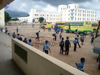 A photograph of students in light blue uniform shirts and dark trousers walking across a dirt yard in front of a large white school building with a blue, yellow and red geometric panel. Several yellow school buses are parked, including one labelled Saraswathi Matric Hr Sec School. Many students carry backpacks, and some hold bags. A few adults are visible in the background. Dark storm clouds hang over distant hills, and a stone wall with a lattice opening runs along the foreground.