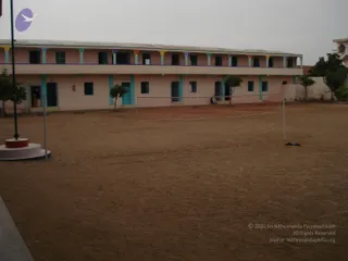 A photograph of an empty dusty courtyard in front of a two-storey pink building with cream and teal colonnaded arches, barred windows and a teal door. A volleyball-style net is strung across the sand on white posts, and a flagpole with a round white and red base stands at left. A small tree is planted near the building, and the sky is overcast. No people are visible in the courtyard except a small distant figure near a doorway at left.