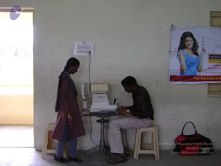 Indoor eye camp scene in a plain whitewashed room with a doorway at left. A young girl in a maroon kurta and dark grey dupatta stands at a table facing a seated young man in a brown jacket and light trousers, who is writing on a clipboard beside an automatic refractor machine. A wall poster at right shows a smiling woman in a blue top holding eyeglasses with the slogan 'For the best in eye care'. Two small stools and a dark red-and-black bag sit on the floor. No a person appearance is visible in the scene.