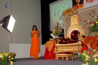 A standing figure in an orange saffron robe with an orange-and-gold beaded mala stands on a stage with palms pressed together in anjali mudra, facing a large ornate golden throne (peetha) with dark red velvet cushions and a golden canopy above. Beside the throne is a wooden book stand, a small brass bowl, a golden footstool, and a microphone on a stand. Red and orange draped tables flank the throne, with bouquets of yellow, pink and red flowers in the foreground and on the right. Behind the stage a projection screen shows a colourful image, and a pattered wall hanging with animal motifs covers the backdrop. A softbox light stands at left. The standing figure appears to be a sannyasi or devotee greeting the empty throne.