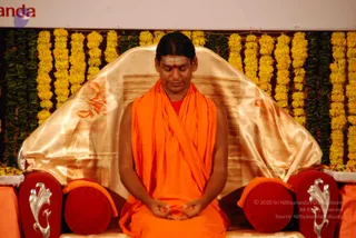 A seated man in an orange saffron shawl over a red undergarment sits in meditation on a gold satin-draped throne with red cushions and silver ornamental armrests. His eyes are closed, hands held in a mudra at his lap, and a red tilak marks his forehead. Behind him hangs a backdrop of yellow marigold garlands and green leaf strings. A watermarked event photo; the figure's shaved-back dark hair and orange robes are visible cues. The appearance is consistent with a person presiding at a meditation session, though identity is uncertain from appearance alone.