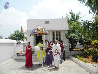 A photograph of a small white ashram-style building with a rounded front door and a barred window, set in a paved courtyard with a flowering shrub and palm trees. A group of about nine people stands in front of it: a woman in a red sari at left holds a metal pot, two children in yellow outfits stand behind her, a woman in a purple sari is at center, and a man in a white dhoti and white shirt holds a bundle of white cloth. A colourful striped umbrella canopy stands over the entrance. A small saffron flag flies on the roof at left.