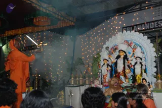 A photograph of a night puja at a decorated pandal. On the left, a figure in an orange saffron robe and orange head covering holds a lit aarti lamp toward the shrine, with smoke drifting through the air. The shrine on the right holds a large white-and-blue arched backdrop framing a deity murti with a multi-armed goddess figure in red and black, flanked by two smaller figures in white and gold, with a lion mount at the base and oil lamps and offerings in front. Strings of warm fairy lights decorate the canopy and walls. The backs of several devotees' heads are visible in the lower foreground. The sign on the right reads SRI NANDES, partly cut off. The aarti-holder's identity is uncertain from appearance alone; the saffron-clad figure is described by role.
