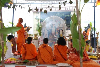 An outdoor puja under a temporary shelter decorated with banana leaves and mango leaf strings. A standing priest in orange robes and an orange head wrap holds a brass lamp, with smoke rising beside him. Several seated participants in orange and maroon cloth sit on an orange mat, and a younger person in white and yellow kneels at left. In front of them is a large deity murti of a goddess with multiple arms, framed by a white lace arch, flanked by two attendant figures. Trays of flower petals, leaves, grains and offerings sit on the ground, and a sign above reads Sri Nanjundeshwara Temple. Brass lamps and a tall lamp stand at right.