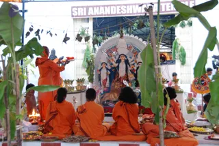 Outdoor puja at Sri Anandeshwari Temple beneath a banner reading Sri Anandeshwari Temple. A deity murti of a goddess with a lion mount stands within a white arched frame, flanked by decorated figures. Several saffron-robed sannyasis sit cross-legged on orange cloth in front, while a standing priest in orange holds a brass plate of offerings and a tall lamp burns in the foreground. Banana leaves and brass pots of flowers frame the scene, with marigolds and fruit on plates before the murti. Visible figures are all in orange robes; no person is clearly identifiable as the a person.