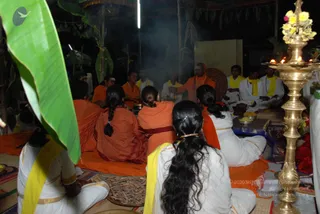 A night-time homa at a temporary open-air pavilion with banana-leaf and palm decorations. Smoke rises from a fire pit at the centre, where a bald man in orange robes and a rudraksha mala sits facing the fire, his hands raised in ritual gesture. Around him, several people in orange robes and white dhotis with yellow shawls sit on mats and cloth. In the foreground, two women with long braided hair in orange saris and a woman in white with a yellow shawl sit with their backs to the camera. On the right, a tall brass lamp with lit wicks and a yellow flower garland stands beside offering plates. A cloth partition and sprinkled leaves complete the setting.