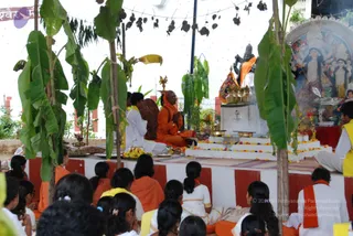 A photograph of a ritual outdoors under a canopy strung with mango leaves and banana plants. On a raised white platform with red-and-white striped sides, a seated figure in orange robes with an orange head covering presses palms together in prayer, with a figure in white beside him. A dark stone deity murti draped in orange cloth stands on a pedestal with brass vessels and marigold garlands, and a framed goddess image with offerings sits to the right. Rows of seated devotees, many in white and orange, face the platform, and a man in a yellow vest kneels at the far right near a brass lamp and flower arrangement. The scene reflects a homa-style puja with floral offerings.