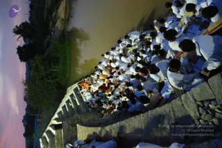 A rotated photograph at dusk shows a large crowd of people dressed in white seated in rows on stone steps beside a water body, many holding books or papers. At the lower left a figure in orange and saffron clothing sits near the front of the group, and several people in white stand and bow in the foreground.