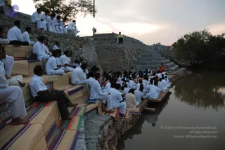A photograph of an outdoor gathering at dusk beside a still pond with stone steps. Many seated people wearing white clothing sit on striped woven mats along the stone embankment and steps, with a few men in white on the upper wall. A figure in saffron-orange robes sits on the stone steps near the water's edge, facing a seated speaker in yellow clothing standing on a raised stone platform at the top of the steps. The pale sky and reflections in the water suggest early evening. The setting resembles a satsang or meditation session held outdoors beside a water tank.