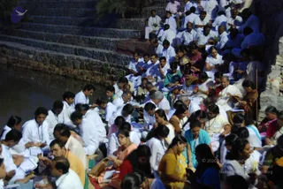 A large outdoor gathering of seated delegates at dusk beside a water body with stone steps. Many people wear white clothing and white lanyard badges, while some women wear saris in yellow, teal, red and pink. Several hold papers, notebooks or small items and appear to be reading or writing. The seated crowd fills the stepped stone embankment and the ground along the water's edge. No individual can be clearly identified as the a person, and no depiction of him is visible.