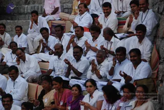 A large outdoor gathering of seated men and women in white clothing, sitting on striped cushions and mats on stone steps. Many men wear white shirts and white dhotis with lanyard name badges and rudraksha-style beaded necklaces, some clapping or gesturing with raised hands. In the foreground several women wear pink, maroon and white saris and white shawls, some with glasses, smiling and clapping. The scene conveys an attentive audience at a meditation therapy session. No single person is identifiable as the central speaker in this frame, and no a person candidate is clearly visible.