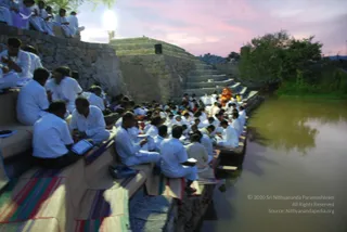 A photograph of a large crowd seated on tiered stone steps beside a pond at dusk, with a pink and blue sky above. Many people wear white clothing and sit on colourful striped mats, some holding books or papers and others with hands pressed together. A small figure in orange robes sits near the top of the steps by the water, with a large lamp glowing on the left wall. Trees and a stone embankment frame the scene.