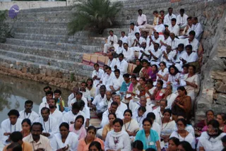 A large outdoor gathering of devotees seated on stone steps beside a water tank at dusk. Dozens of people, mostly dressed in white with blue lanyard badges, sit on striped mats in rows, many with hands pressed together in namaskar. Some women wear saffron, pink, teal and turquoise saris and salwar, and a few carry papers. A small palm tree grows on the upper terrace, and the stone wall rises behind the crowd. No a person candidate is visible among the seated participants.