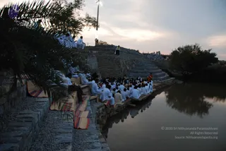 A large group of people dressed in white sits in rows along stone steps and a stone embankment beside a still water tank at dusk. Many wear white shirts and white dhotis, and some sit on striped woven mats laid across the stone ledges. Palm fronds hang in the foreground on the left. Behind the seated rows, a figure in orange stands on a raised stone platform near a lamp post, and stone steps rise on the right under a pale evening sky. The water reflects the seated crowd and trees.