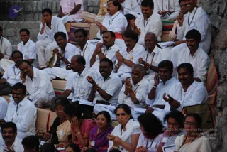 A seated audience of men and women at an outdoor session, sitting on stone steps and cushioned mats along a rocky wall. Most men wear white shirts and white dhotis or trousers, with white lanyard-style ID badges and some with rudraksha-like beaded necklaces; several clap or gesture with raised hands. Women wear saris in pink, purple and white and bangles. Papers and notebooks rest on laps. No clearly identifiable a person figure is visible.