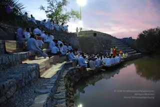 Dusk outdoor gathering at a stone-terraced pond with stepped stone seating. Many people in white clothes sit in rows on mats along the terraces and the water's edge, facing a figure in orange robes standing on the steps at right, with a small group in white near him. A tall floodlight glows at the top of the frame against a pink and lavender evening sky, and palm fronds frame the left edge. The pond reflects the seated crowd. No identification of the orange-robed figure is made from appearance.