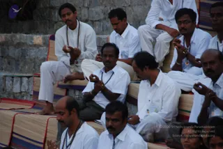A photograph of a seated group of men in white shirts and white trousers on striped cushioned mats on stone steps, several wearing rudraksha malas. Many have hands held in mudra-like gestures. A stone retaining wall forms the backdrop.