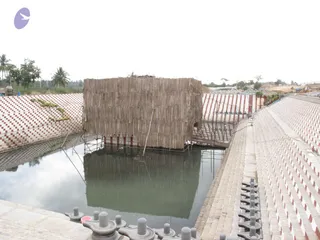 A photograph of a construction site showing a large square structure wrapped in bamboo matting or woven reed screens, standing in the middle of a rectangular water-filled reservoir. Stepped embankments on both sides are faced with red and white striped brick or tile, and a row of grey stone shivalinga-style lingams with pedestals lines the bottom edge in the foreground. Scaffolding poles and wooden planks surround the structure, and a sandy area with construction machinery and stacked rubble sits in the background under an overcast sky with palm trees. No people are visible.